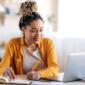 Focused stylish female student studying remotely from home, using a laptop, taking notes on notepad during online lesson, e-learning concept, smiling