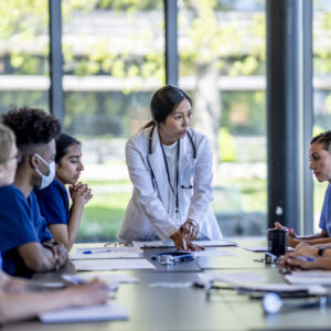 A small group of medical student residents gather around a boardroom table to to meet with their medical team lead. They each have cases out on front of them as they work together collaboratively to discuss each one. They are each dressed professionally in medical scrubs and are listening attentively to the doctor leading the meeting at the head of the table.